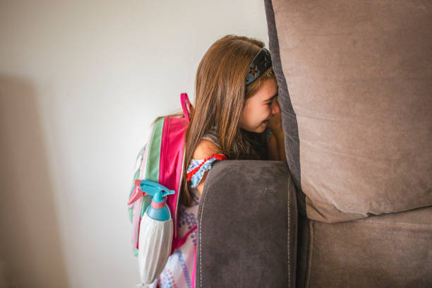 Scared school girl refuses to go to school, and makes it known by hiding behind the sofa Displeased school girl, showing her fear and negative emotional, while hiding and and refusing to go to school backpack sprayer stock pictures, royalty-free photos & images