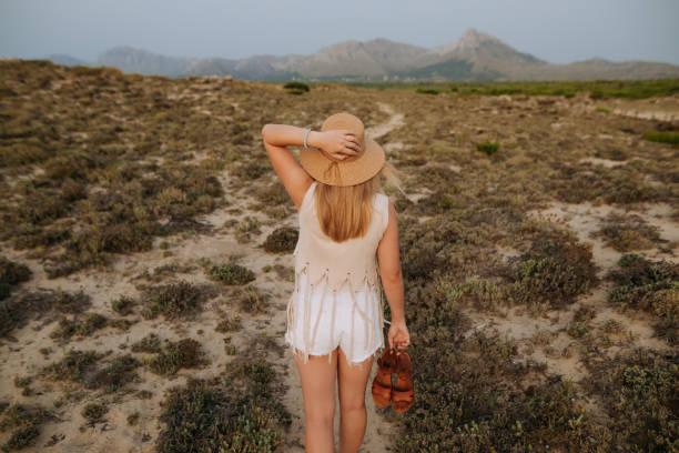 istock Blond young woman with a summer hat and dressed in boho clothes from behind standing between the green dunes in Majorca and a beautiful mountain landscape far in the background 1330579986