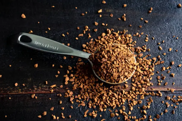 A measuring spoon filled with instant coffee granules that are spilling over onto a wooden table