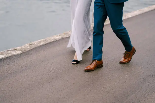 Photo of Legs of the groom in blue trousers and the bride in a white dress are walking along the asphalt road over the sea. Close-up