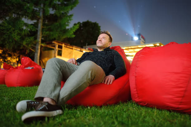 homem bonito deitado no travesseiro na grama e assistindo filme no cinema ao ar livre em parque público. passar a noite perfeita ou o fim de semana ao ar livre. lazer e entretenimento para as pessoas no verão. - night in the city - fotografias e filmes do acervo