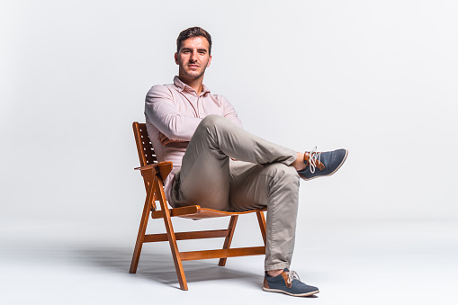 Portrait of a confident young adult businessman wearing a light pink shirt and beige pants. Smiling and sitting on a chair against white background.