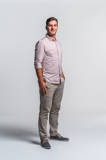 Portrait of a confident young adult businessman wearing a light pink shirt and beige pants. \nHands on pockets standing against white background.