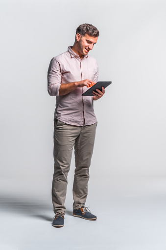 Portrait of a confident young adult businessman wearing a light pink shirt and beige pants. using a digital tablet standing against white background.
