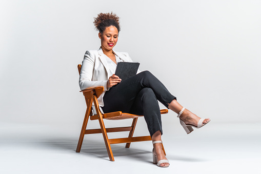 Portrait of Confident Mixed race woman with curly hair in white blazer, white shirt and black pants holding a digital tablet sitting on a chair against white background.