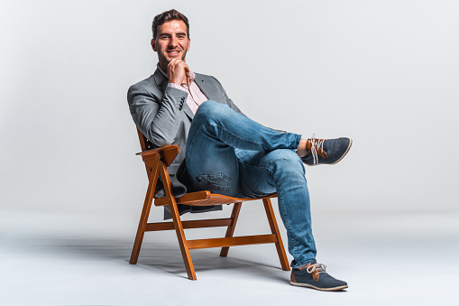 Portrait of a confident smiling young adult businessman in smart casual clothes sitting on a chair using laptop against white background.