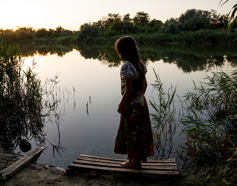 A girl in a long colored sundress stands on the shore of the lake and looks into the water at sunset. Rear view.