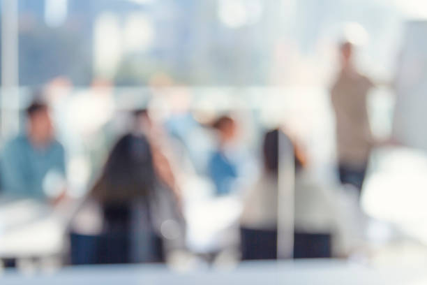 Defocussed image of Business people watching a presentation. A mature man is writing on the whiteboard with charts and graphs. They are sitting in a board room, All are casually dressed.