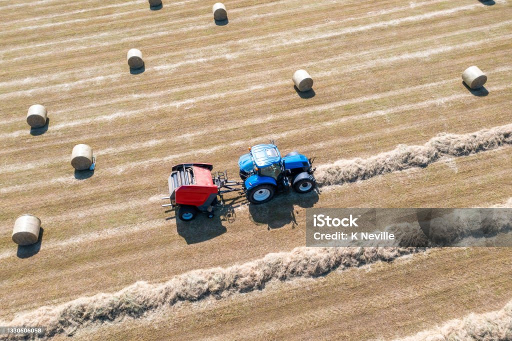 Tractor and hay baler at work A drone point of view, looking down at a farmer driving a blue tractor, that is towing red a hay baler. Lines of dried cut grass, are being fed into the baler and being made into large round bales of hay, that will be used as winter feed for livestock. There are beautiful patterns of harvesting that can only be seen from above. Bale Stock Photo