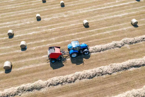 A drone point of view, looking down at a farmer driving a blue tractor, that is towing red a hay baler. Lines of dried cut grass, are being fed into the baler and being made into large round bales of hay, that will be used as winter feed for livestock. There are beautiful patterns of harvesting that can only be seen from above.