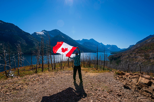 Young girl in a canoe from behind holding canadian flag spreading her arms, surrounded by turqouise blue lake Moraine and mountains