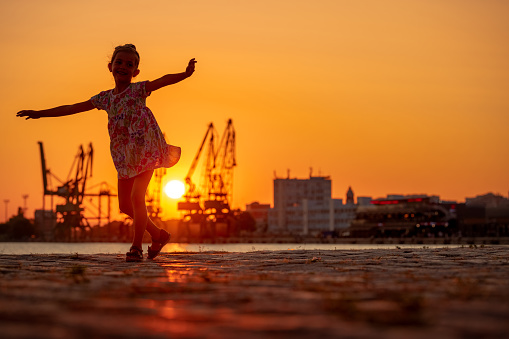 Little girl dancing during sea sunset. Silhouttes of industrial cranes on sea port Varna, Bulgaria