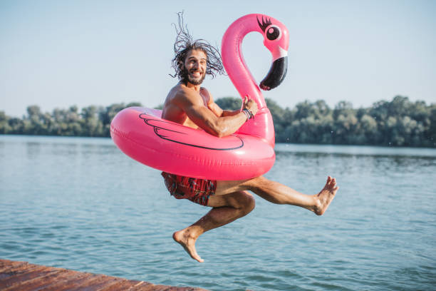 Summer by the river Young man jumping in river from deck. He is jumping with pink flamingo swimming float. summer fun stock pictures, royalty-free photos & images