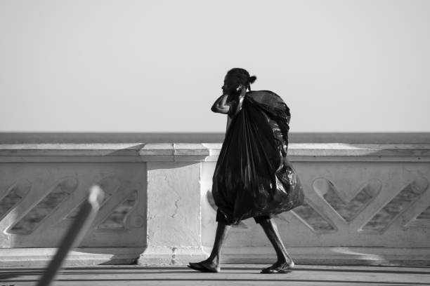 Homeless woman walking Salvador, Bahia, Brazil - October 02, 2020: Homeless woman walking with a bag on her back on the edge of Farol da Barra beach. She looks for beer and soda cans for recycling. walking beach women sadness stock pictures, royalty-free photos & images