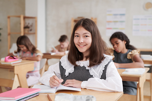 Portrait of smiling schoolgirl wearing uniform while sitting at desk in classroom with wooden decor, copy space