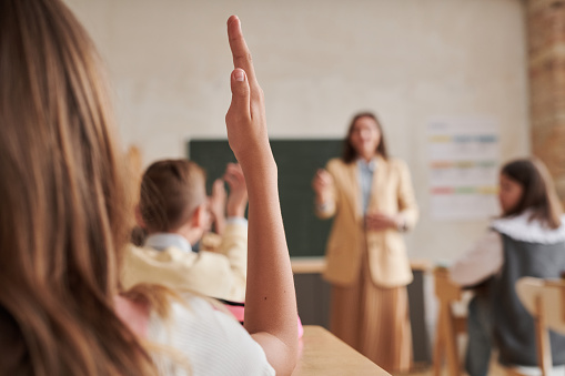 Close up of unrecognizable schoolgirl raising hand in class with female teacher in background, copy space