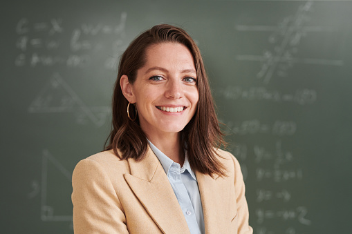 Portrait of young female teacher looking at camera and smiling while standing by blackboard in school classroom, copy space