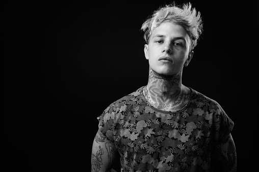Studio shot of young handsome rebellious man with blond hair and tattoos against black background in black and white