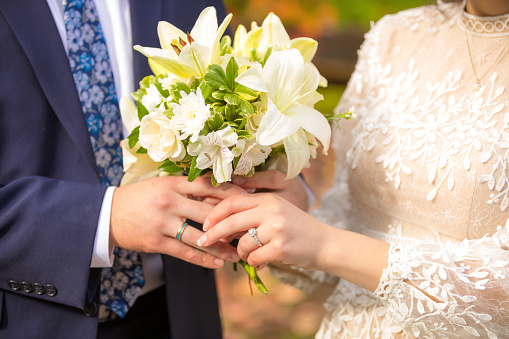 Hands of young newly married couple holding flower bouquet of white lilies, showing wedding rings