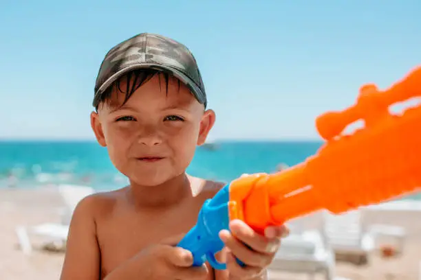 A child boy stands near the sea with a water gun close-up portrait. Water games of children near the water on the beach. High quality photo