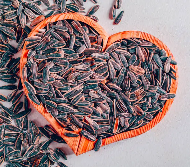 Black sunflower seeds on heart shaped board and white background, top view.
