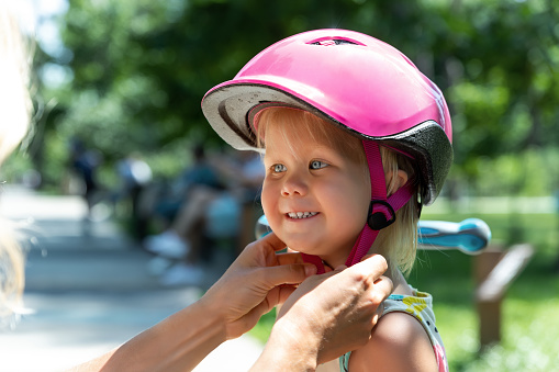 Close-up mom parent hand put on and fasten safety helmet on cute blond caucasian toddler girl for riding bike or scooter city street park outdoors on summer day. Child sport activity protection care.