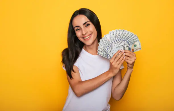 Young happy excited beautiful woman emotionally looking in the camera with a lot of money in her hands and having fun isolated on yellow background