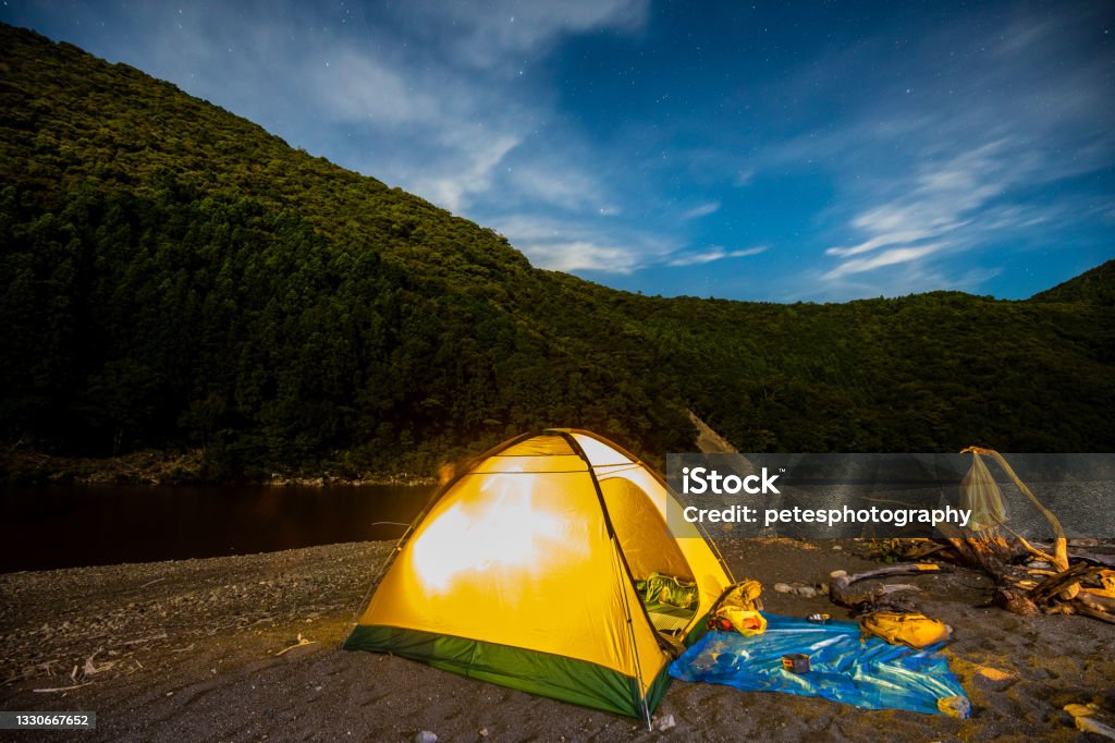Illuminated tent camping under the stars by a lake A tent illuminated on a sandy bank by a river with hills behind under a starry night sky. Kumano - Mie Stock Photo