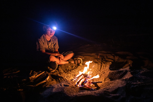 A young mixed race boy by his camp fire he made in a sandy pit with his head lamp on.