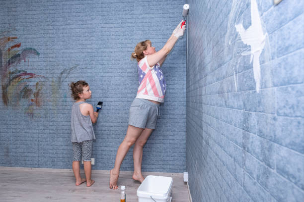 famiglia di caucasici. una giovane donna che tiene in mano un rullo di vernice e una bambina stanno facendo lavori di ristrutturazione della stanza di un bambino. una donna applica lo sgattoiore per carta da parati al muro. - paint roller copy space action adult foto e immagini stock