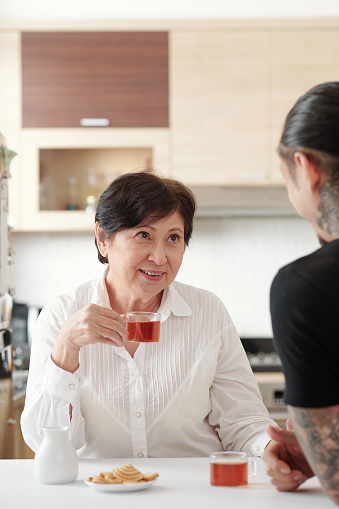 Asian mature woman sitting at the table drinking tea and talking to her son in the kitchen