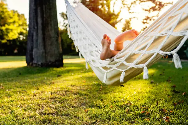 Cute little feet of barefoot child sticking out of hammock in backyard garden at sunset. Child summer holidays healthy lifestyle and outdoors leisure activity. Vacation at home, slow living, gadget detox