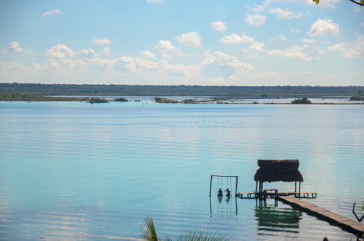 Panorama view of a couple in a swing in Bacalar lagoon in Mexico. Honeymoon destination in the Caribbean. Swings inside an exotic lake. Tropical summer vacations. Tranquility and paradise background.