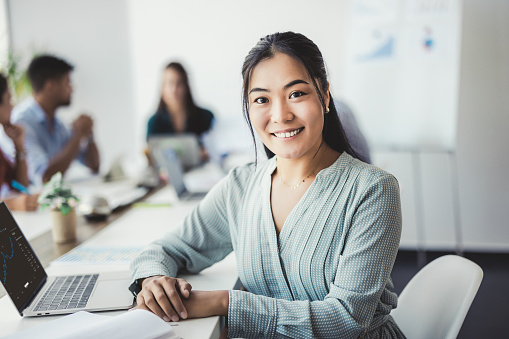 Portrait of young smiling asian woman looking at camera while her colleagues working in the background. Shallow focus.