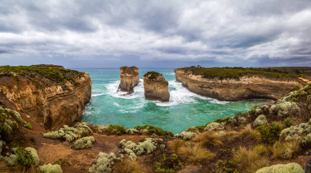 a wide panorama from the rocky brown shore to the turquoise ocean, rocky islands and the dark sky at ard gorge, island arch and razorback, australia. - the loch ard gorge imagens e fotografias de stock