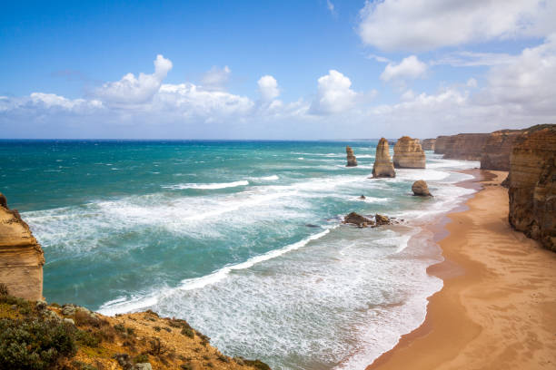 pintoresca tarjeta de fotos panorámicas en impresionantes vistas de las olas del océano, la playa de arena naranja y los acantilados escarpados bajo el cielo azul los doce apóstoles, great ocean road, victoria - twelve apostles sea rocks fotos fotografías e imágenes de stock