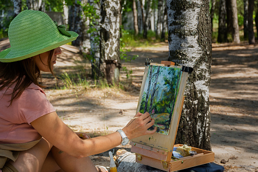 A young woman draws plein air. An artist in a hat in a birch forest writes a sketch from nature. The concept of creativity, favorite hobby, hobby. The process of painting a picture with acrylic paints