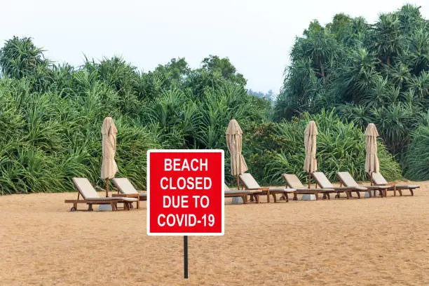 Photo of ''Beach closed due to COVID-19'' warning sign against loungers and folded beach umbrellas in a desolate tropical beach