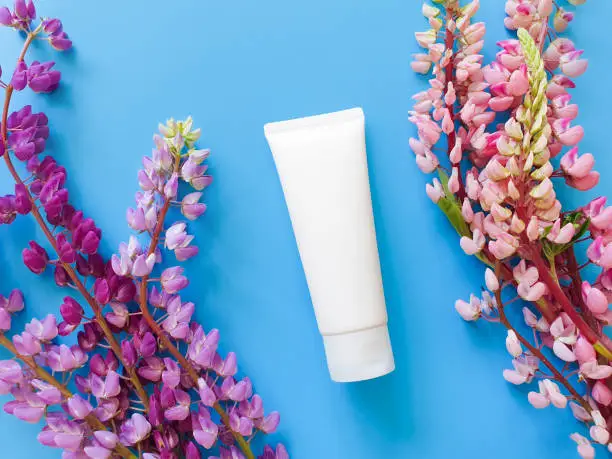 Photo of Mockup of white squeeze bottle plastic tube and lupines flowers on blue table. Template, top view. Natural organic spa cosmetics concept.