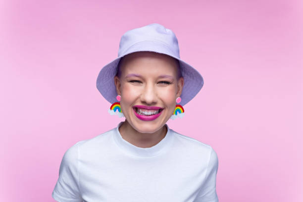 Headshot of woman with rainbow earrings Excited young woman wearing white t-shirt, lilac bucket hat and funny rainbow earrings laughing at camera. Studio portrait on pink background. bucket hat stock pictures, royalty-free photos & images