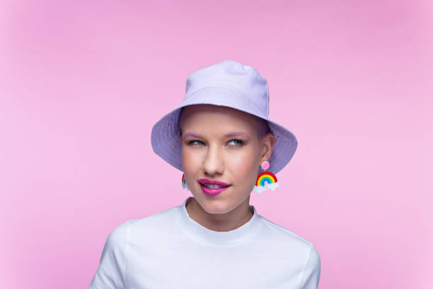 Headshot of woman with rainbow earrings Thoughtful young woman wearing white t-shirt, lilac bucket hat and funny rainbow earrings looking away. Studio portrait on pink background. bucket hat stock pictures, royalty-free photos & images