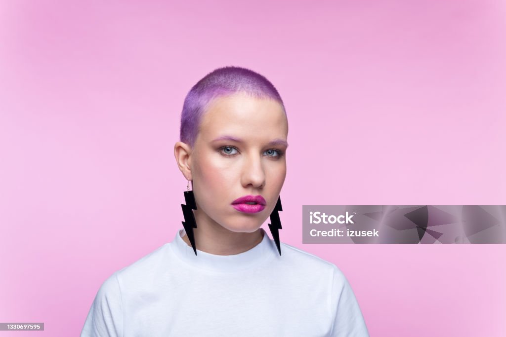Headshot of woman with short purple hair Young woman wearing white t-shirt and big earrings looking at camera. Studio portrait on pink background. Lightning Stock Photo