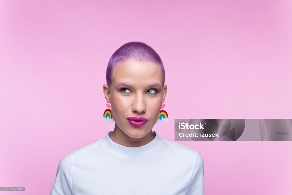 Headshot of woman with short purple hair and rainbow earrings Thoughtful young woman wearing white t-shirt and funny rainbow earrings looking away. Studio portrait on pink background. 20-29 Years Stock Photo