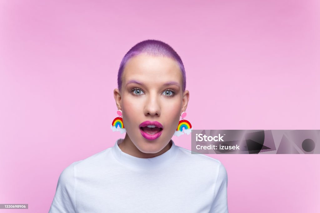 Headshot of woman with short purple hair and rainbow earrings Surprised young woman wearing white t-shirt and funny rainbow earrings staring at camera. Studio portrait on pink background. Surprise Stock Photo