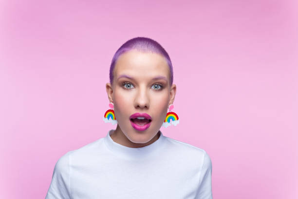 Headshot of woman with short purple hair and rainbow earrings Surprised young woman wearing white t-shirt and funny rainbow earrings staring at camera. Studio portrait on pink background. purple hair stock pictures, royalty-free photos & images