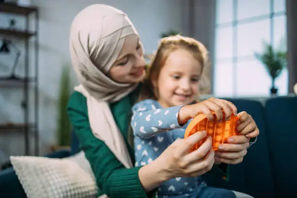 Photo of Muslim woman in hijab, sitting on sofa at home and playing with pop it fidget toy with her daughter