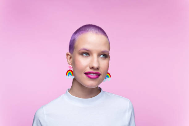 Headshot of woman with short purple hair and rainbow earrings Thoughtful young woman wearing white t-shirt and funny rainbow earrings looking away. Studio portrait on pink background. transgender stock pictures, royalty-free photos & images