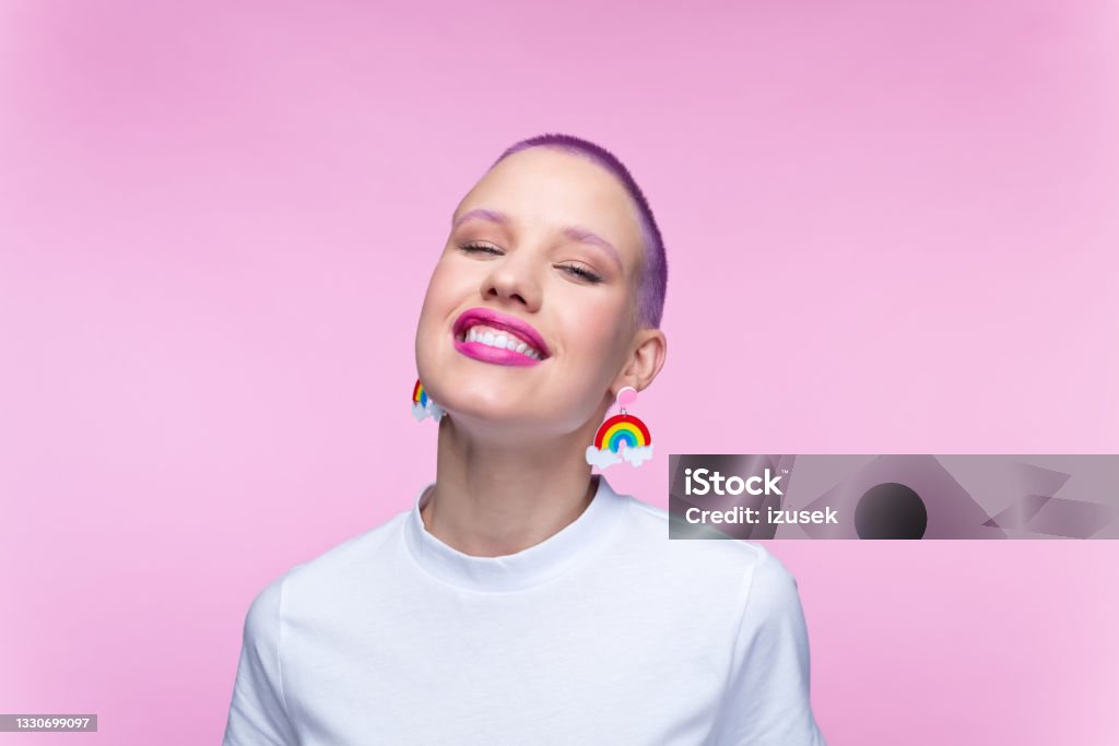 Headshot of woman with short purple hair and rainbow earrings Cheerful young woman wearing white t-shirt and funny rainbow earrings smiling at camera. Studio portrait on pink background. Transgender Person Stock Photo