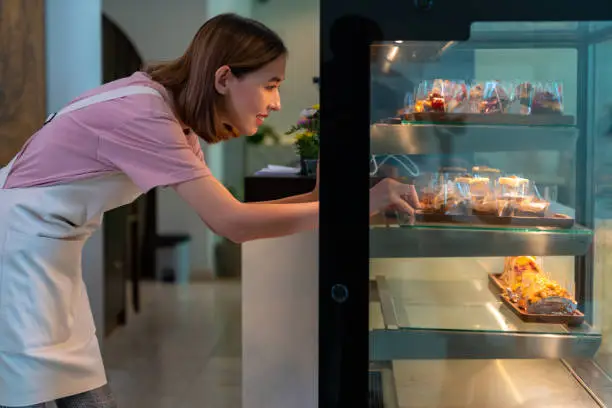 Photo of Asian woman coffee shop employee placing cake in bakery refrigerator