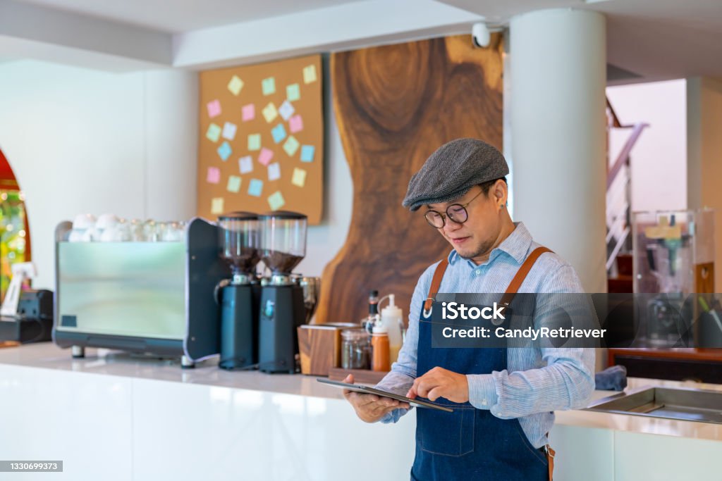 Asian man coffee shop owner checking inventory on digital tablet Asian man coffee shop manager walking behind bar counter working on digital tablet checking and counting inventory for ordering product for cafe. Small business restaurant owner entrepreneur concept Small Business Stock Photo
